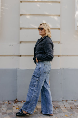 A woman stands in profile against a white building wall with shutters, wearing a dark textured long-sleeve top, wide-leg blue jeans, and black flats.