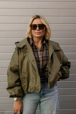 A woman wearing a green hooded bomber jacket over a brown and blue plaid shirt, paired with light-wash blue jeans and dark sunglasses, standing against a gray horizontal siding wall.