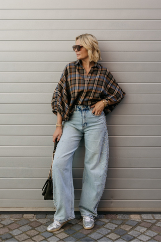 A woman stands against a large gray garage door wearing a brown and black plaid flannel shirt, light-wash wide-leg jeans, white sneakers, and sunglasses, holding a black handbag.