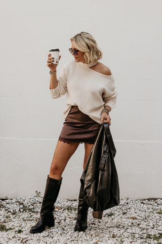 A woman stands against a white wall on a gravel surface, wearing an off-the-shoulder cream sweater, a brown mini skirt, and black knee-high boots. She holds a coffee cup in one hand and a black jacket in the other.