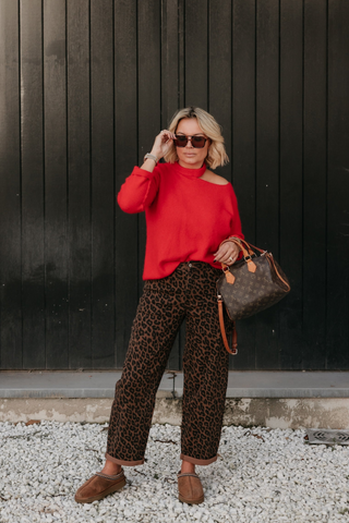 A woman stands in front of a large black metal door framed by brick walls, wearing a bright red off-the-shoulder top, leopard-print wide-leg pants, brown mule shoes, and holding a brown monogram handbag.
