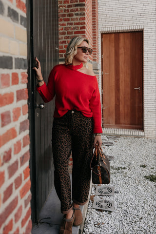 A woman wearing a bright red off-the-shoulder long-sleeve top and leopard-print pants stands in an outdoor alleyway next to a brick wall, holding a brown leather handbag.