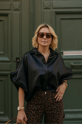 A woman with blonde hair and sunglasses stands in front of a dark green paneled door, wearing a black satin-like blouse with dramatic draped sleeves and leopard-print pants.