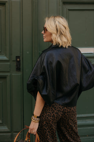 A woman with blonde hair is shown from behind wearing a black leather-like blouse and leopard print pants, standing in front of a dark green paneled door.
