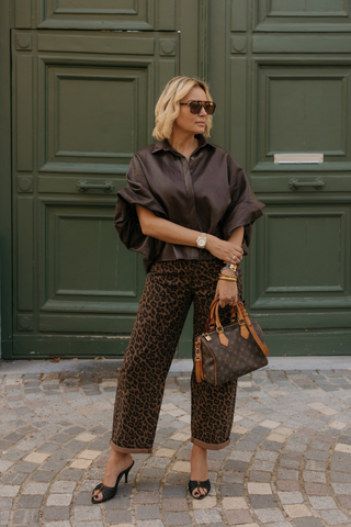 A woman stands in front of large dark green wooden doors wearing a dark brown satin blouse, leopard-print cropped pants, black slingback heels, and holding a brown monogram handbag.
