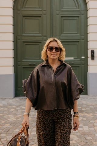 A woman stands in front of a large green arched door wearing a dark brown short-sleeve button-up shirt and leopard-print pants, holding a brown handbag.
