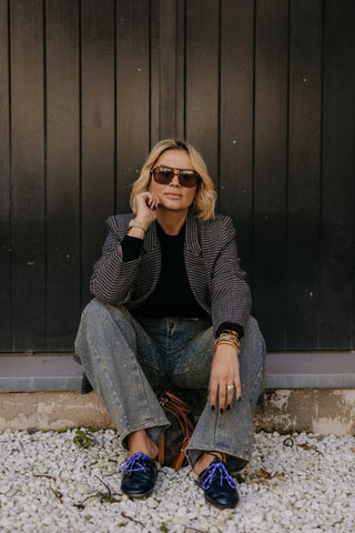 A woman wearing a black and gray houndstooth blazer, black top, wide-leg gray jeans, and dark loafers with bright blue laces sits on a concrete ledge against a dark wood-paneled wall.